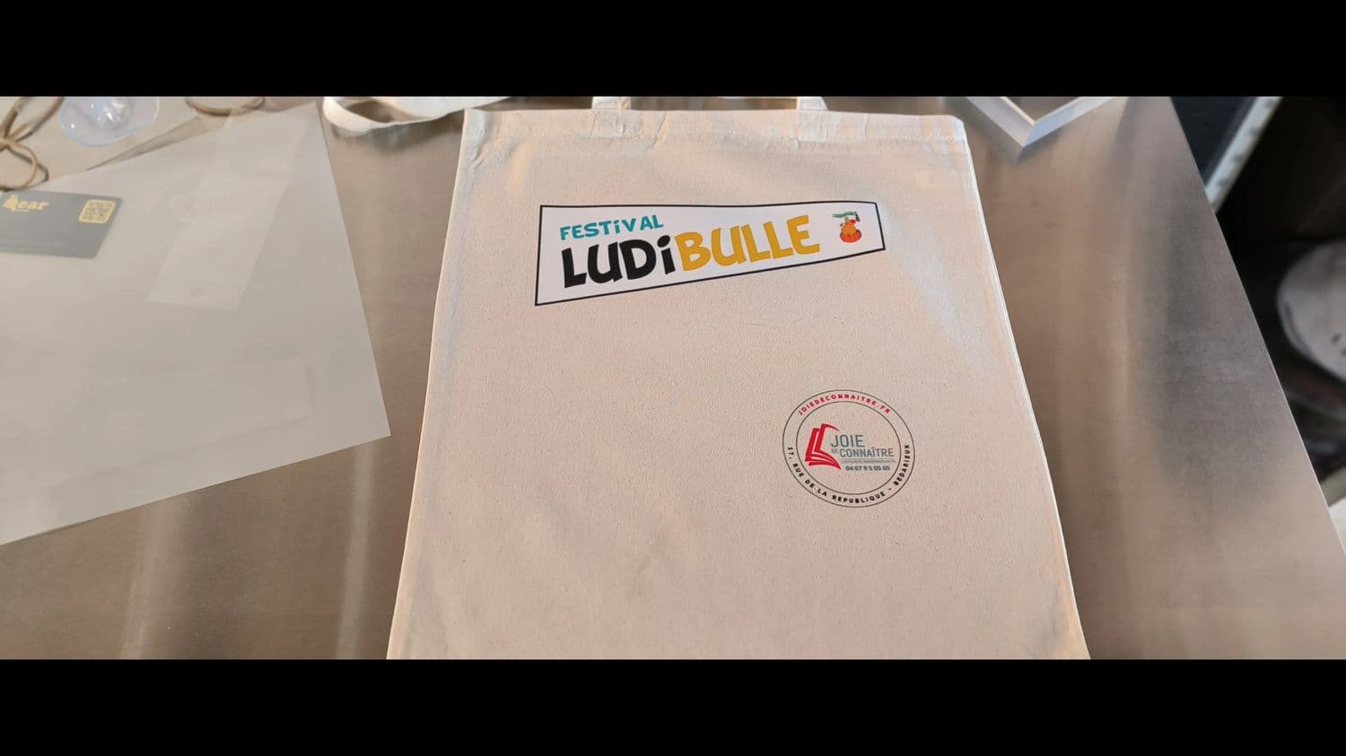 Tote bag coton écru personnalisé : logo du festival LudiBulle en haut, tampon de la librairie Joie de Connaître en bas